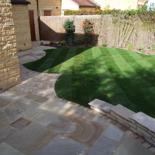 Indian sandstone paving with a low Cotswold stone wall and a sculpted lush green lawn border by rustic fencing and trellis with plants and perennials