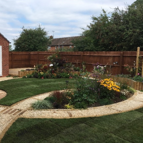 A circular lawn bordered by a low wooden bed of shrubs and perennials, with a winding limestone and cobble path dissecting a lush green lawn and feature plant beds full of colourful shrubs and perennials