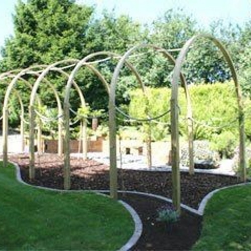 A wooden rose arch walkway over a winding bark path defined by cobble and brick borders next to a lush green lawn and plant beds full of shrubs and perennials