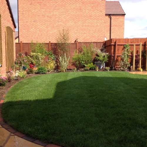 A lush green lawn bordered by colourful plants and perennials and edged with neat brick blocks, and a Cotswold stone paved patio