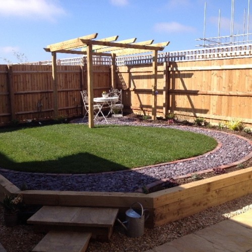 Wooden steps up to a raised path of blue slate shingle and cobbles, surrounding a lush lawn with a wooden pergola creating shade over a private seating area in the corner of the garden