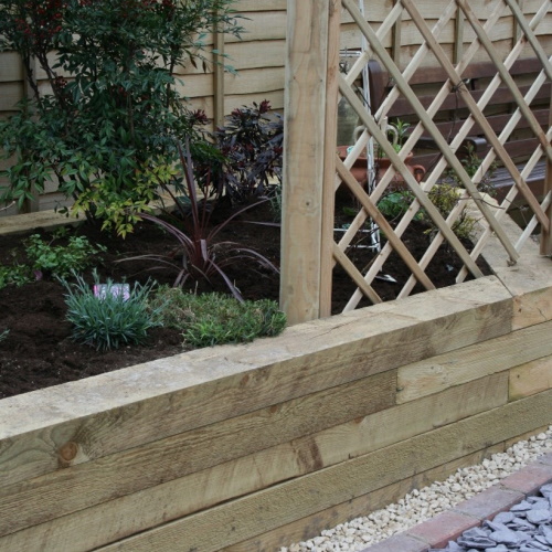 A raised wooden bed packed full of shrubs and grasses with a trellis to grow climbers up and bordered by a limestone, cobble and blue slate shingle path