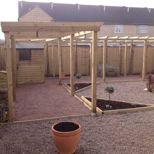 A wooden pergola arch covering a structural garden with angular wooden beds and planted with ornamental trees